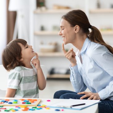 Speech training for kids. Professional woman training with little boy at cabinet, teaching right articulation exercises Speech training for kids. Professional woman specialist training with little boy at cabinet, teaching him right articulation exercises, side view